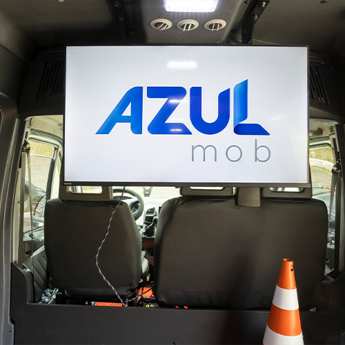 Interior de uma van com tela exibindo “AZUL MOB”, dois assentos em primeiro plano e um cone de trânsito laranja.