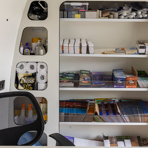 Prateleiras embutidas em compartimento branco armazenando itens como papel higiênico, recipientes e livros.