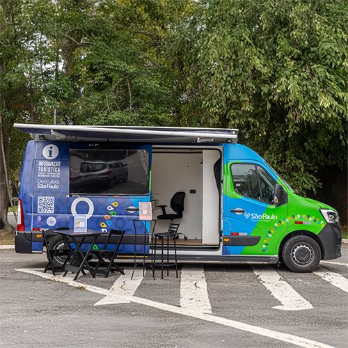 Furgão de serviço móvel azul e verde com porta lateral aberta, mesas e cadeiras, estacionado na rua.