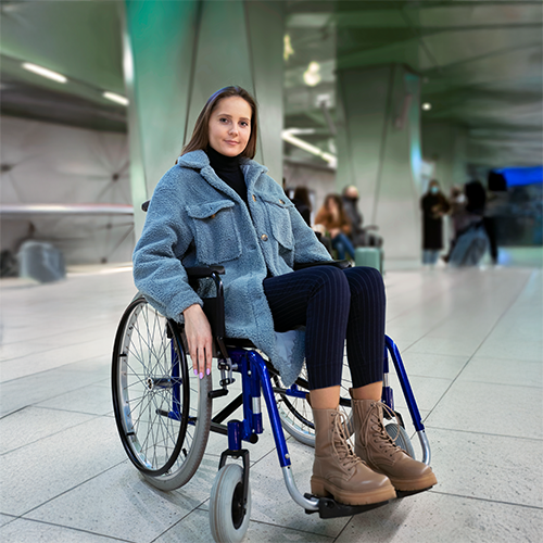 Mulher em cadeira de rodas, vestindo casaco e botas azuis, em um aeroporto.