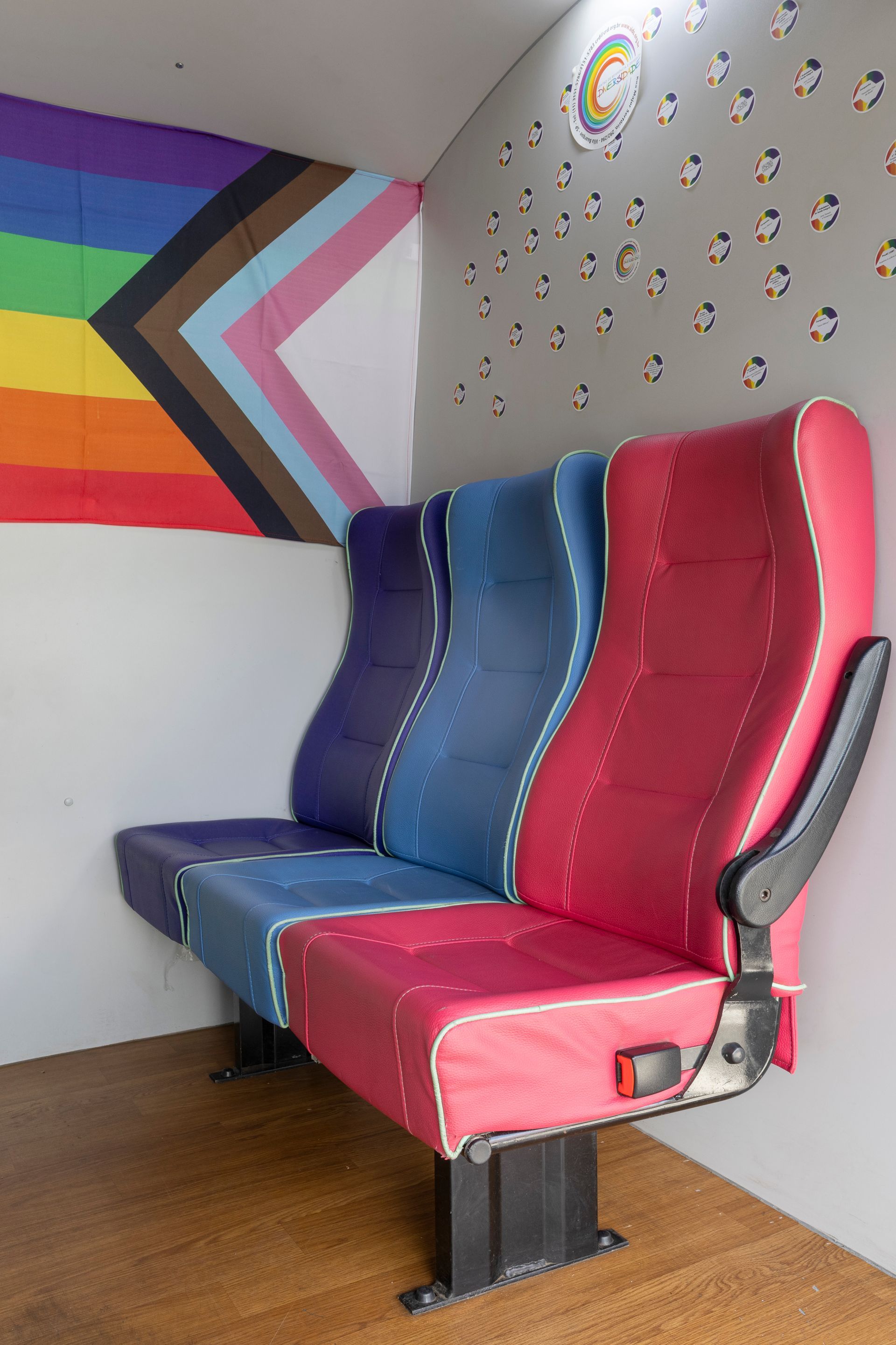Três assentos de ônibus coloridos em um canto, com uma bandeira do Orgulho LGBTQIA+ e pequenos desenhos nas paredes.