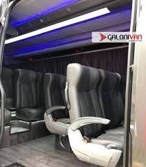 Vista interna de uma van de passageiros com bancos de couro cinza e iluminação interna.