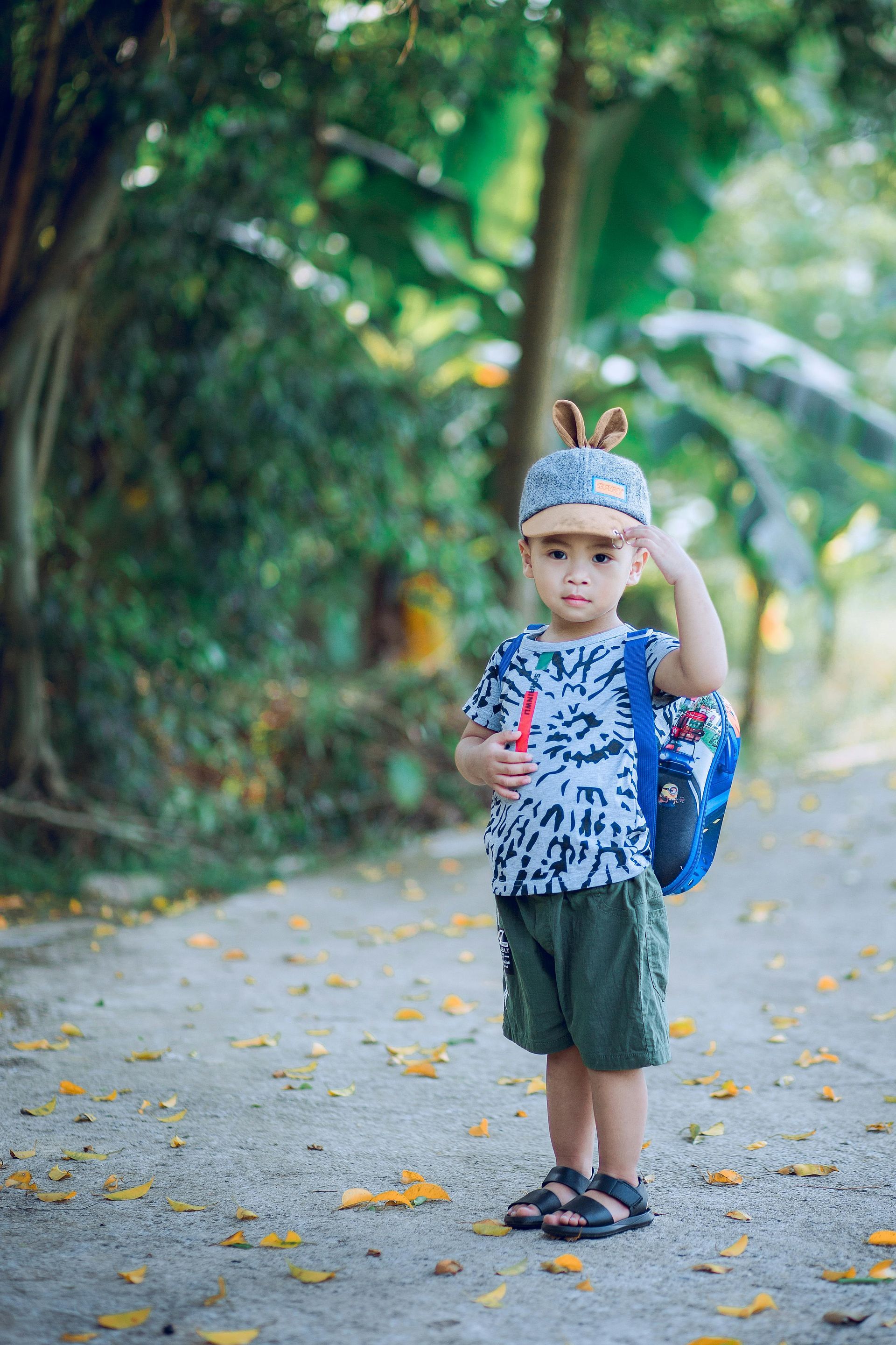 Menino de chapéu azul e mochila, em pé ao ar livre em uma trilha, segurando um objeto vermelho.