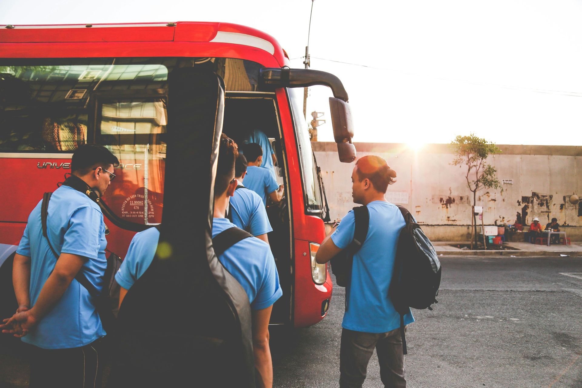 Pessoas vestindo camisas azuis embarcando em um ônibus vermelho, um estojo de guitarra em primeiro plano, luz solar intensa.