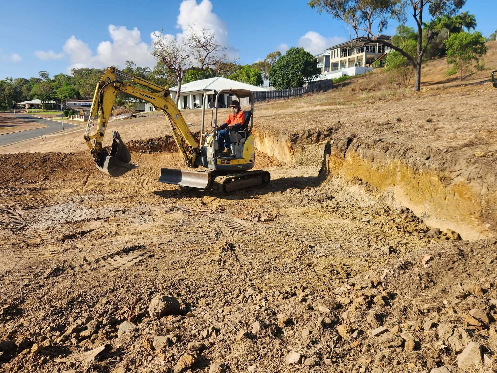 Yellow excavator digging a trench in dirt at a construction site. Operator inside. Trees and houses in the background. . - DJ Brazil earthmoving, truck and tilt tray hire, Bowen QLD