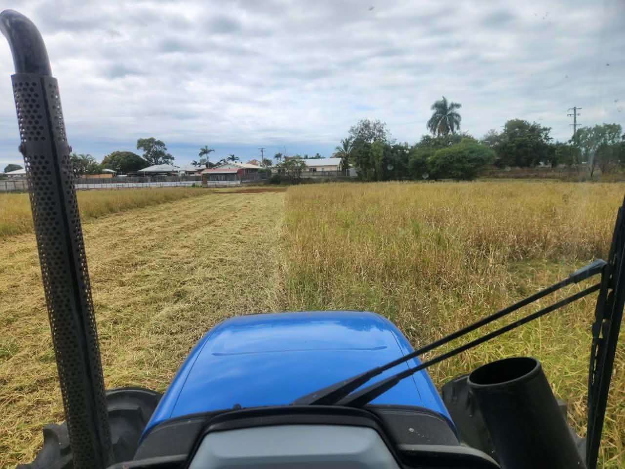 Blue tractor cutting through field; cloudy sky, buildings in background. . - DJ Brazil earthmoving, truck and tilt tray hire, Bowen QLD