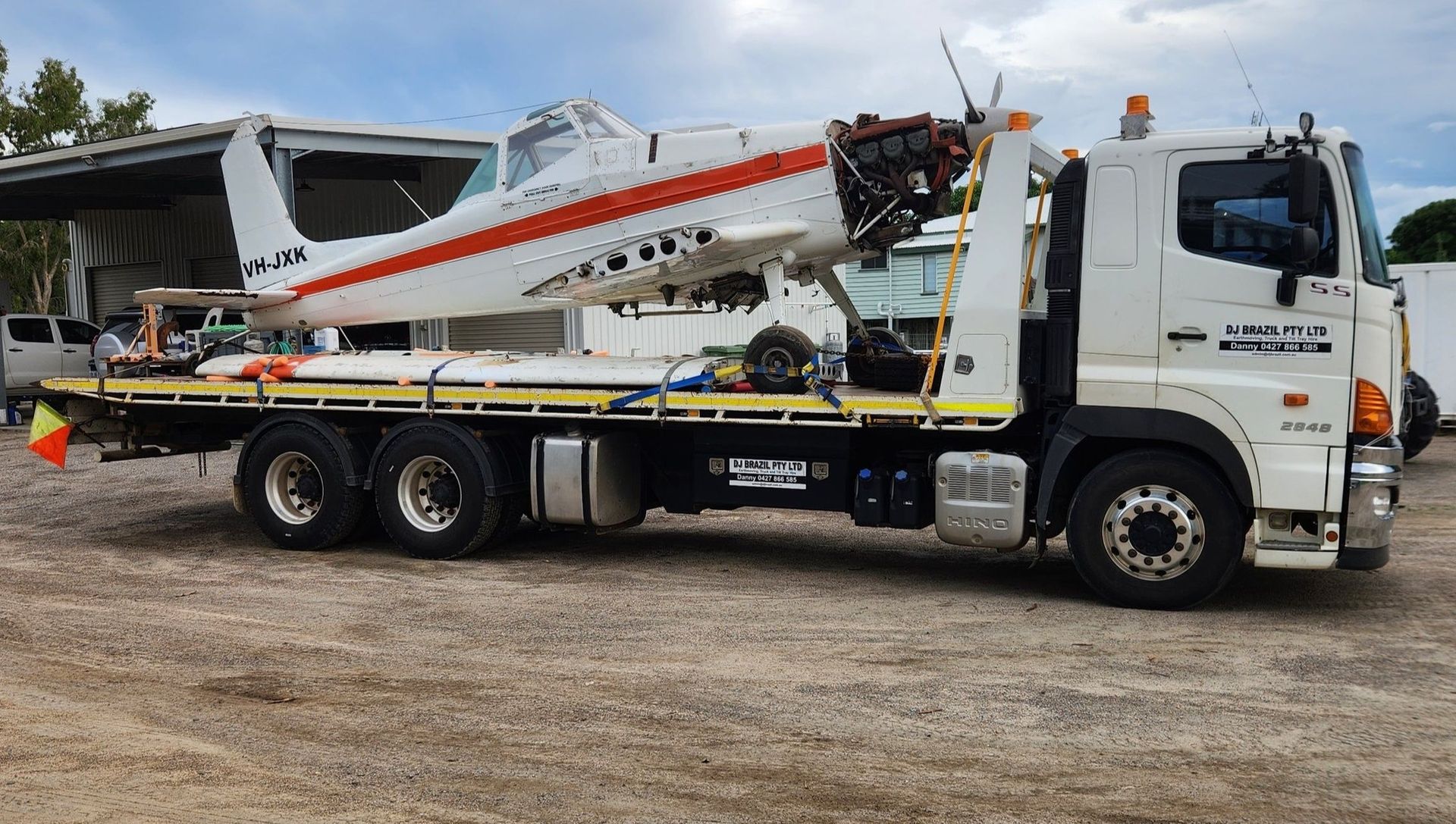 Airplane on a flatbed tow truck. White plane with red stripe, engine removed. Parked outside. - DJ Brazil earthmoving, truck and tilt tray hire, Bowen QLD
