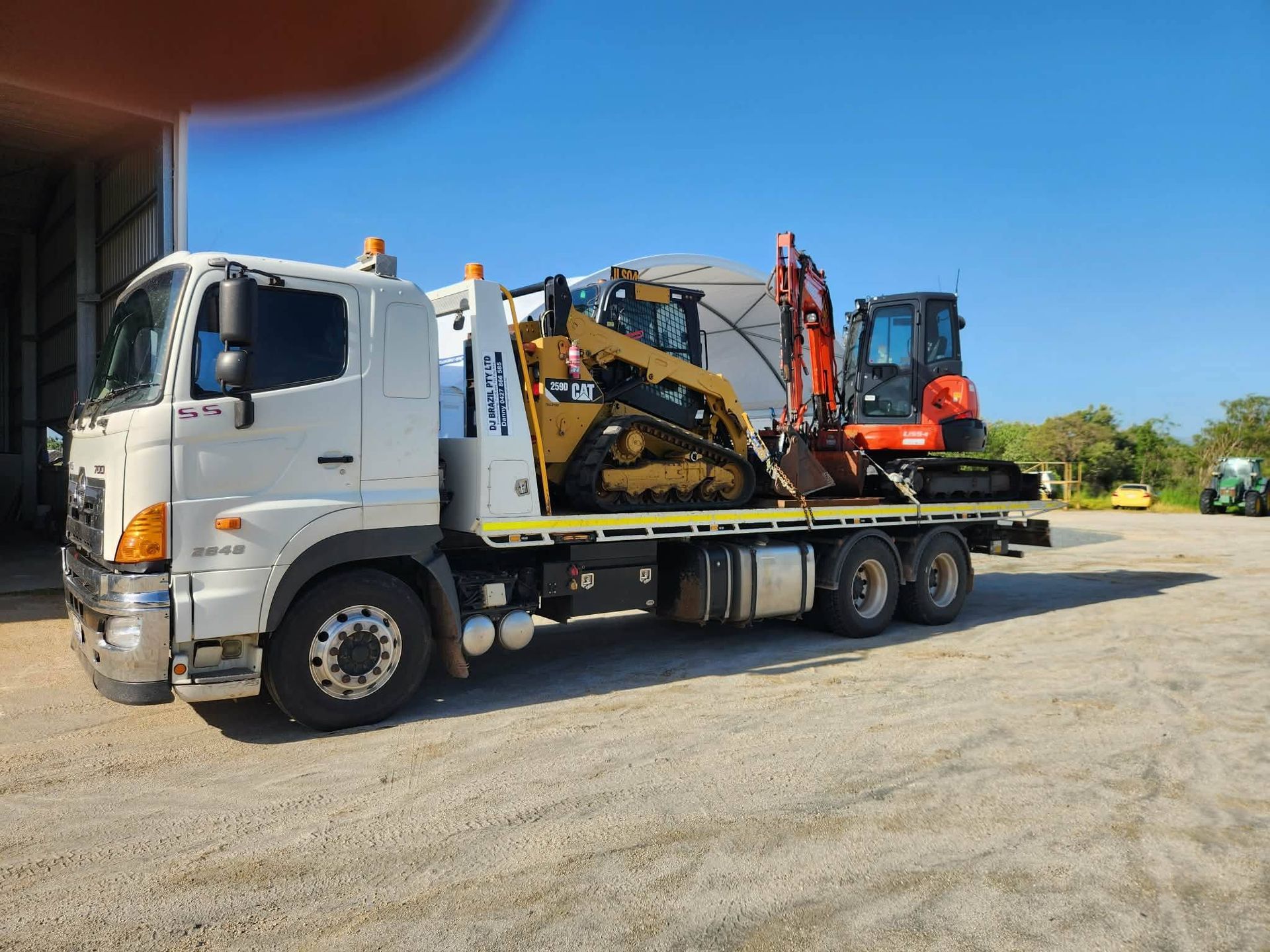 White truck transporting a Caterpillar skid steer and a Kubota excavator on a sunny day. . - DJ Brazil earthmoving, truck and tilt tray hire, Bowen QLD