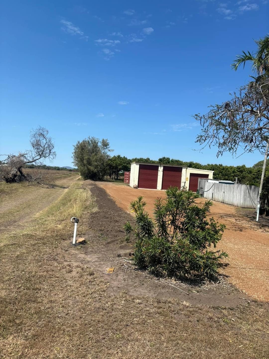 Brown and red garage building with two bays, set in a dry, grassy landscape under a blue sky. . - DJ Brazil earthmoving, truck and tilt tray hire, Bowen QLD