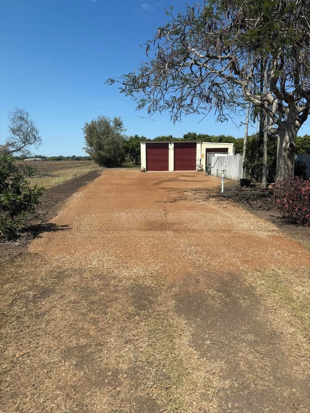 Driveway leading to a two-car garage with red doors, surrounded by trees and shrubs, under a blue sky. . - DJ Brazil earthmoving, truck and tilt tray hire, Bowen QLD