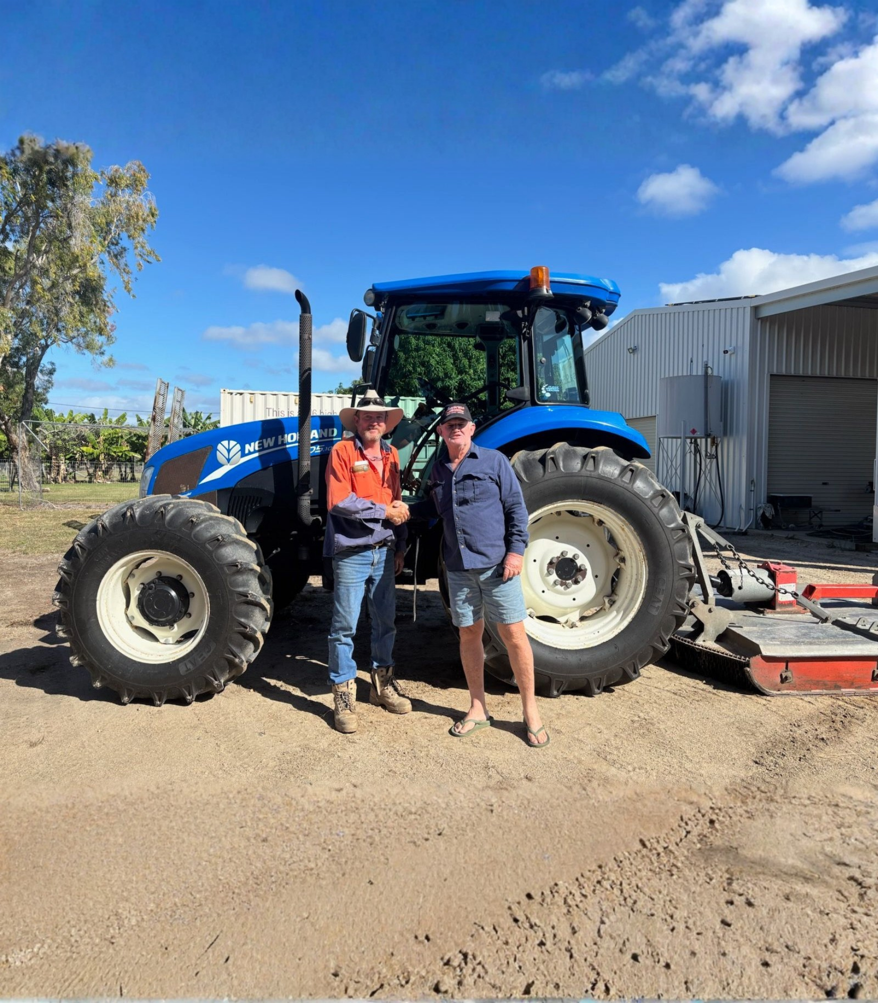 Two men shake hands in front of a blue New Holland tractor on a sunny day. . - DJ Brazil earthmoving, truck and tilt tray hire, Bowen QLD