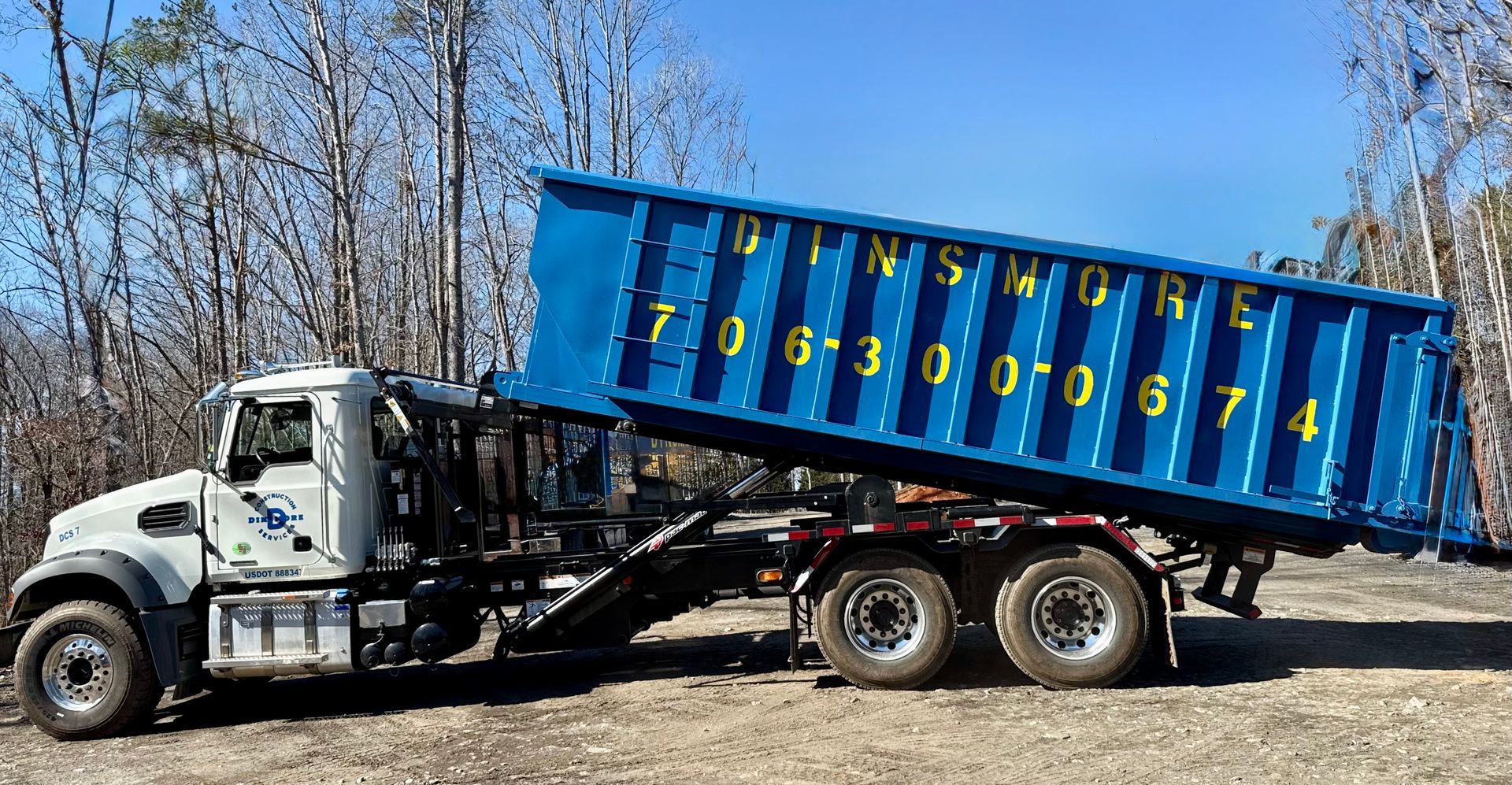 Blue dumpster on a truck, with phone number on the side, on a dirt road.
