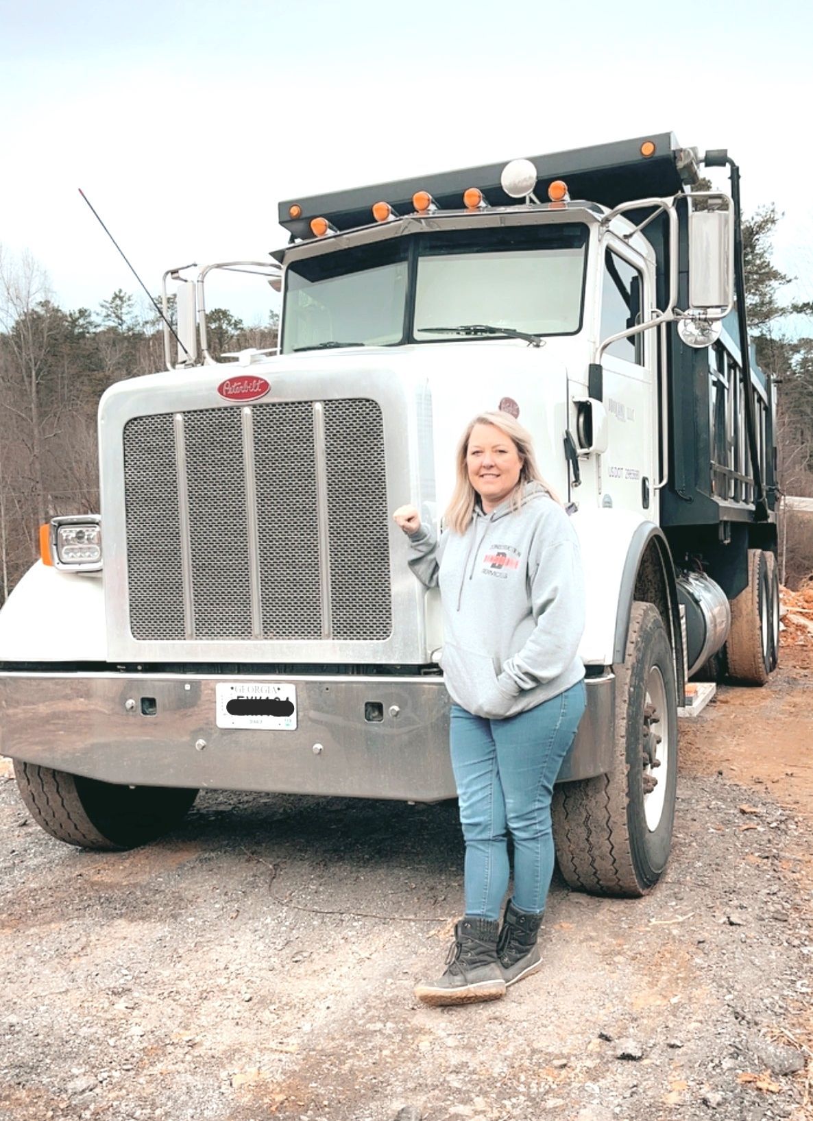 Woman standing next to a white dump truck, smiling. Outdoor setting, overcast sky.