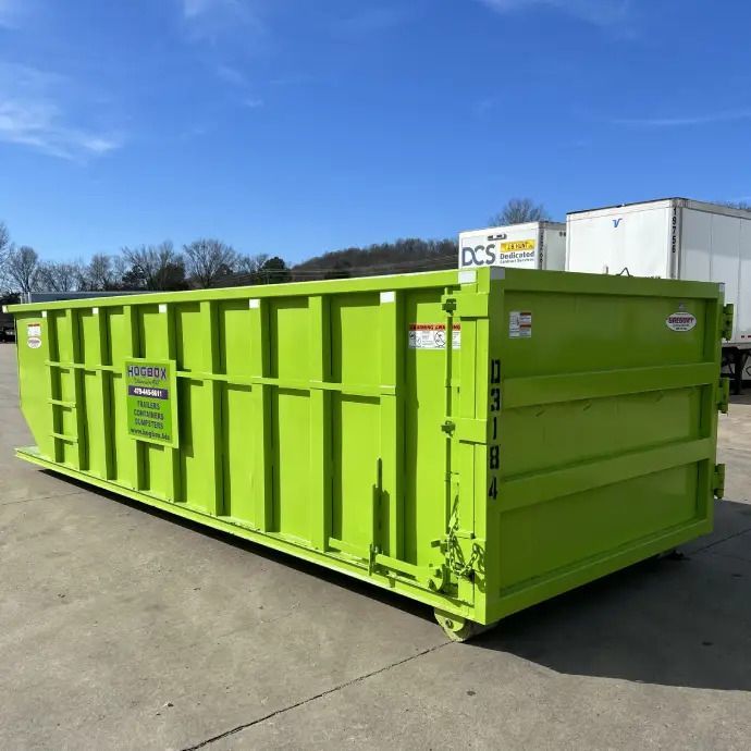 Bright green dumpster in a paved lot under a blue sky, with trees and trucks in the background.
