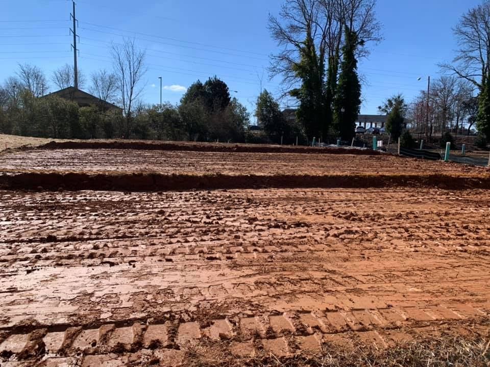Dirt field with tractor tracks and leveled areas, blue sky, trees in background.
