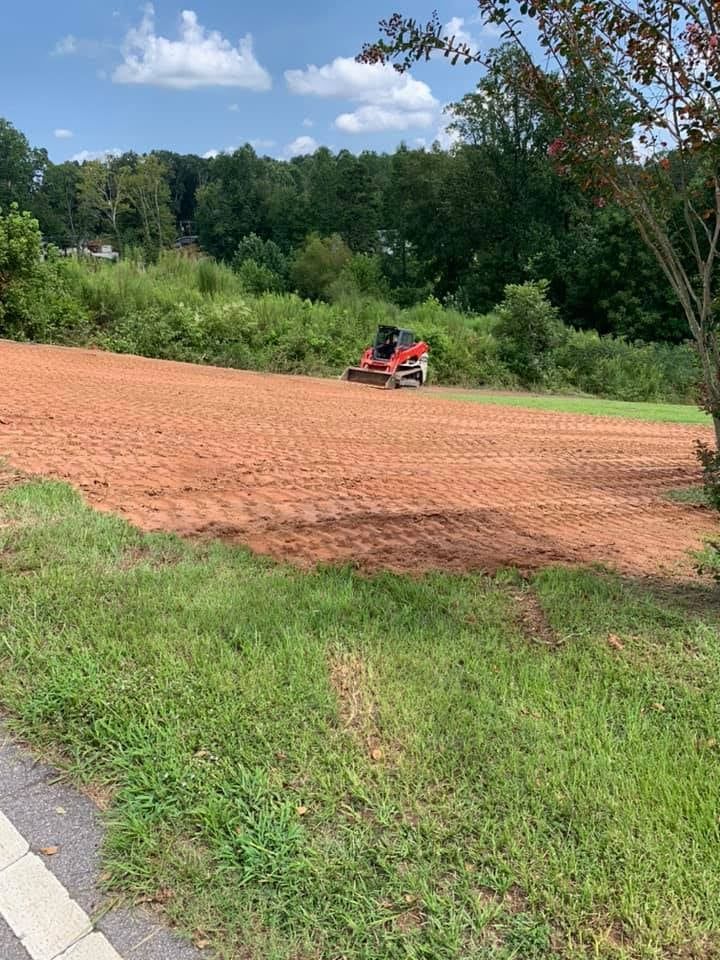 Red soil graded by a small construction vehicle; green grass in foreground, trees and blue sky in background.