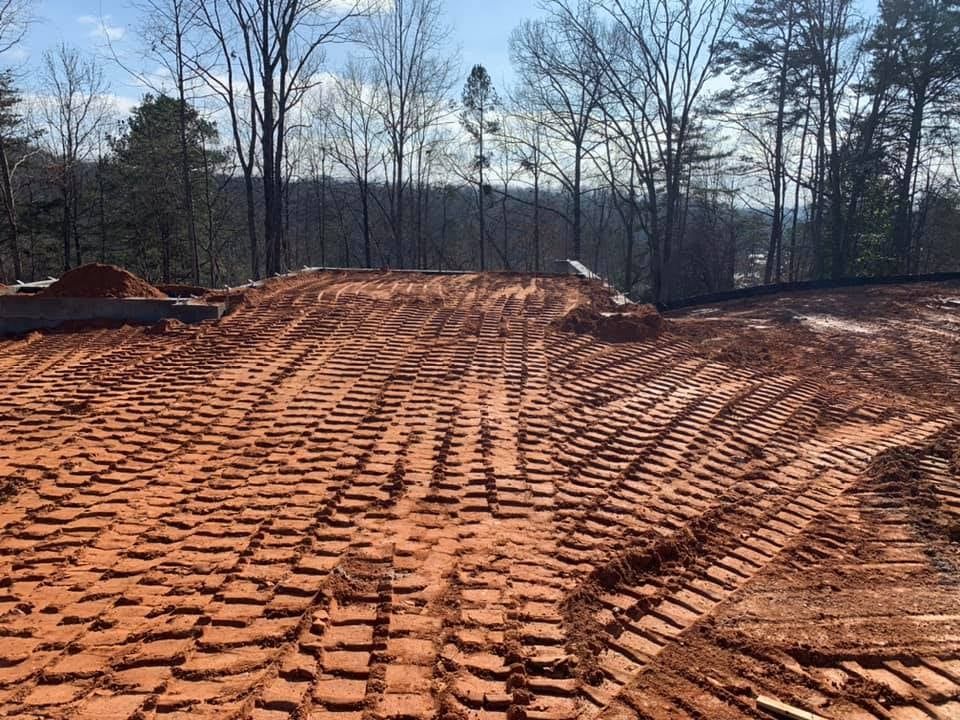 Construction site with red dirt, tire tracks, and a small mound. Trees in the background.