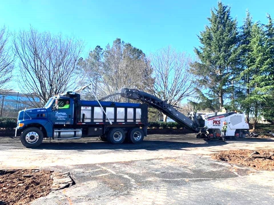 Blue truck and milling machine removing asphalt from a parking lot on a sunny day.