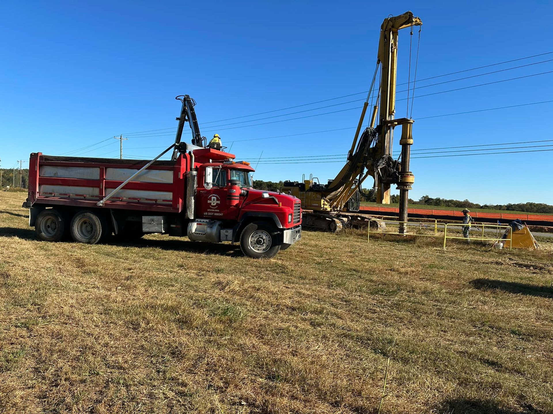 Red dump truck next to a construction site with a drilling machine under a blue sky.