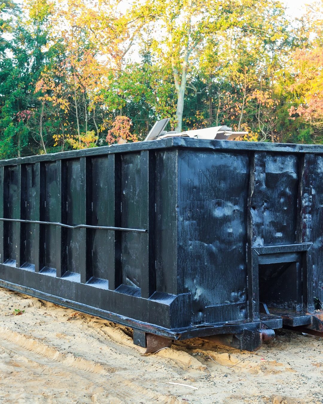 Black dumpster outdoors with fall foliage background.