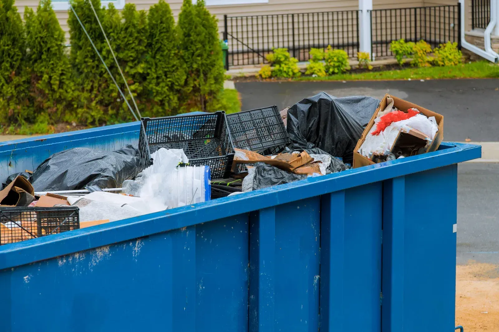 A large blue dumpster filled with black trash bags, cardboard boxes, and plastic crates sits outdoors by a building.
