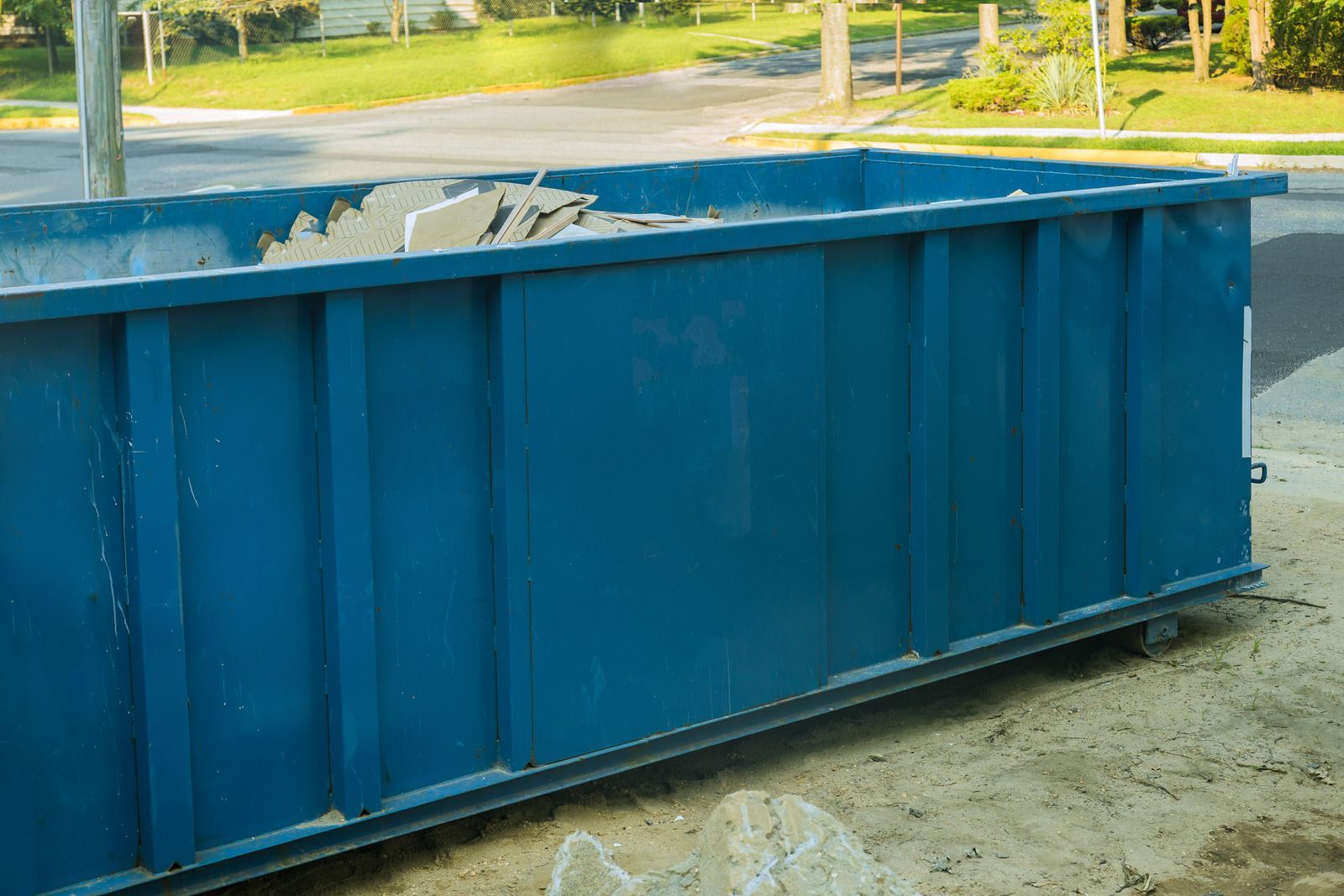Blue dumpster filled with debris, sits beside a street in daylight.