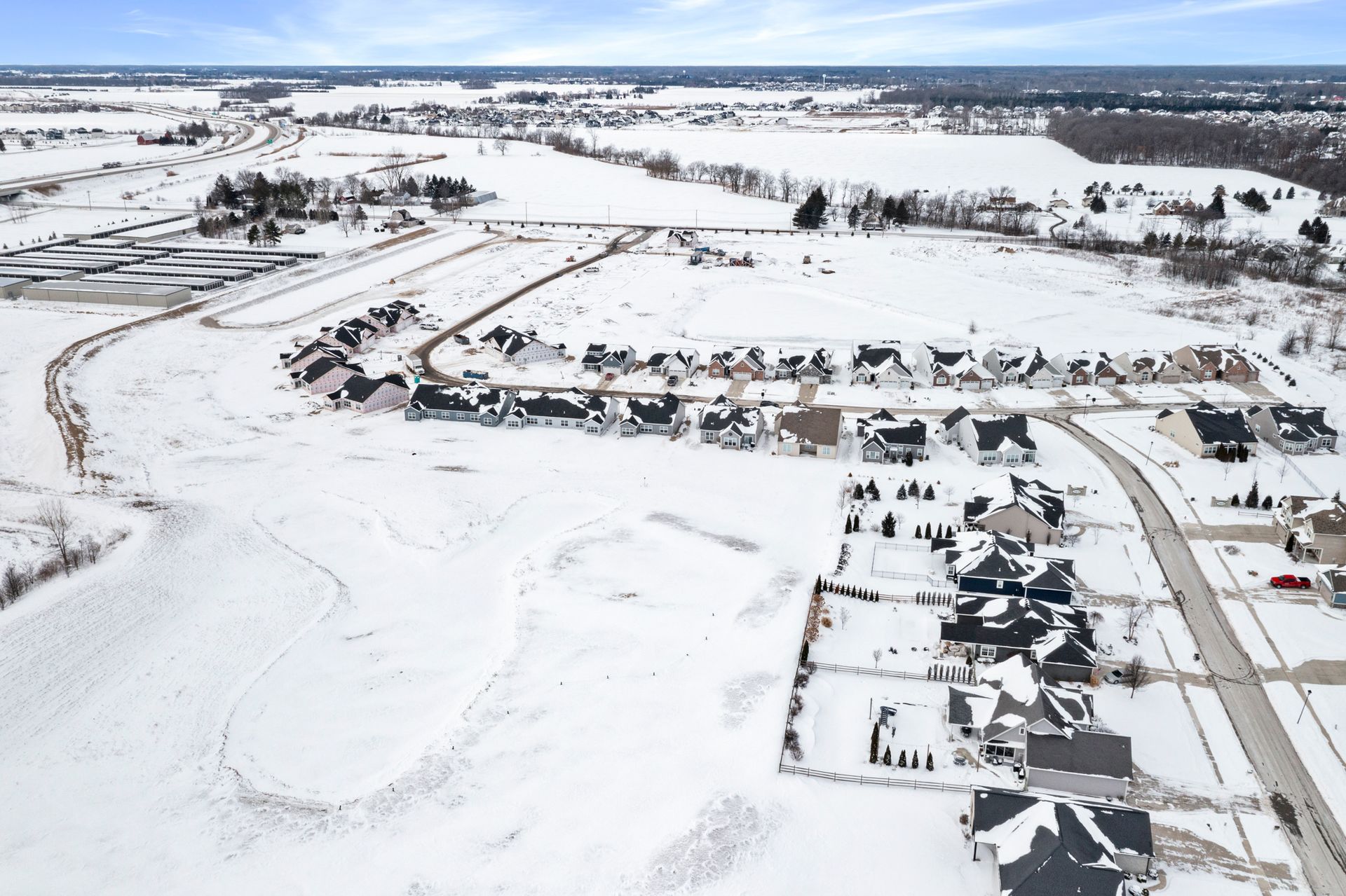 Snowy aerial view of a suburban neighborhood, rows of houses, roads, and open fields, under a cloudy sky.