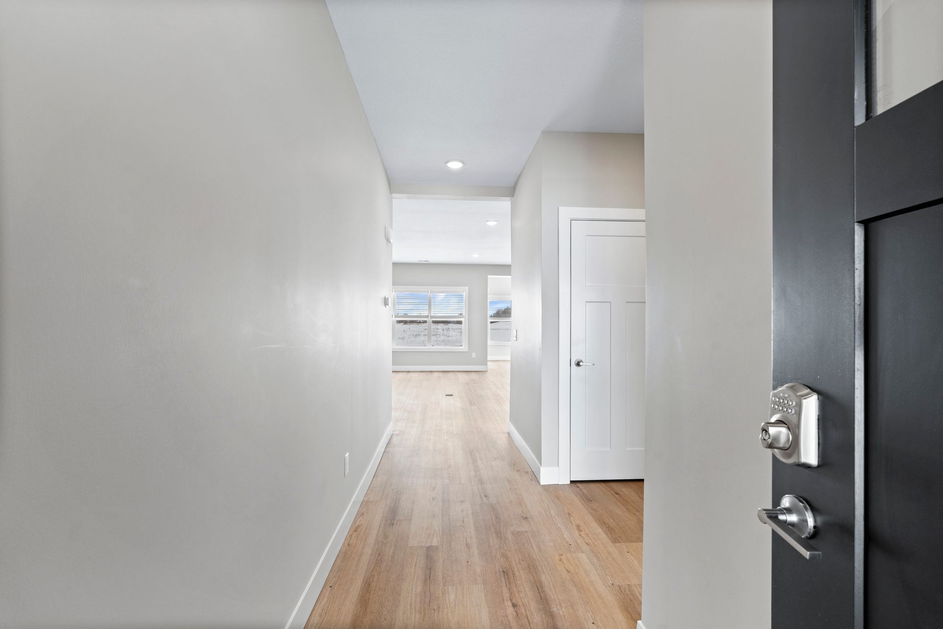 Hallway with light wood floors, leading to a room with windows; open door to right.