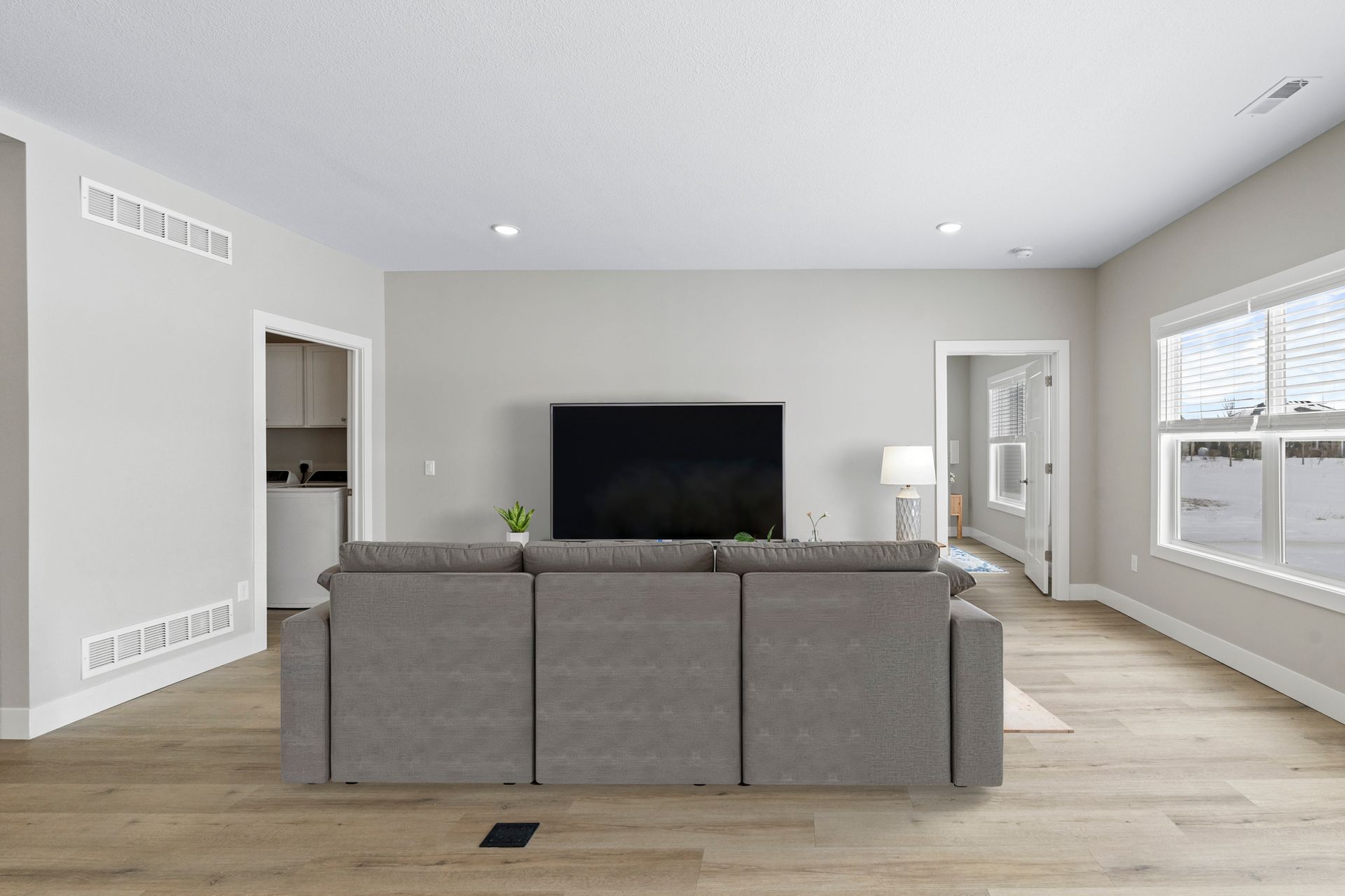 Living room with gray sofa facing a TV on a gray wall. Two windows on right side. Light wood floors.