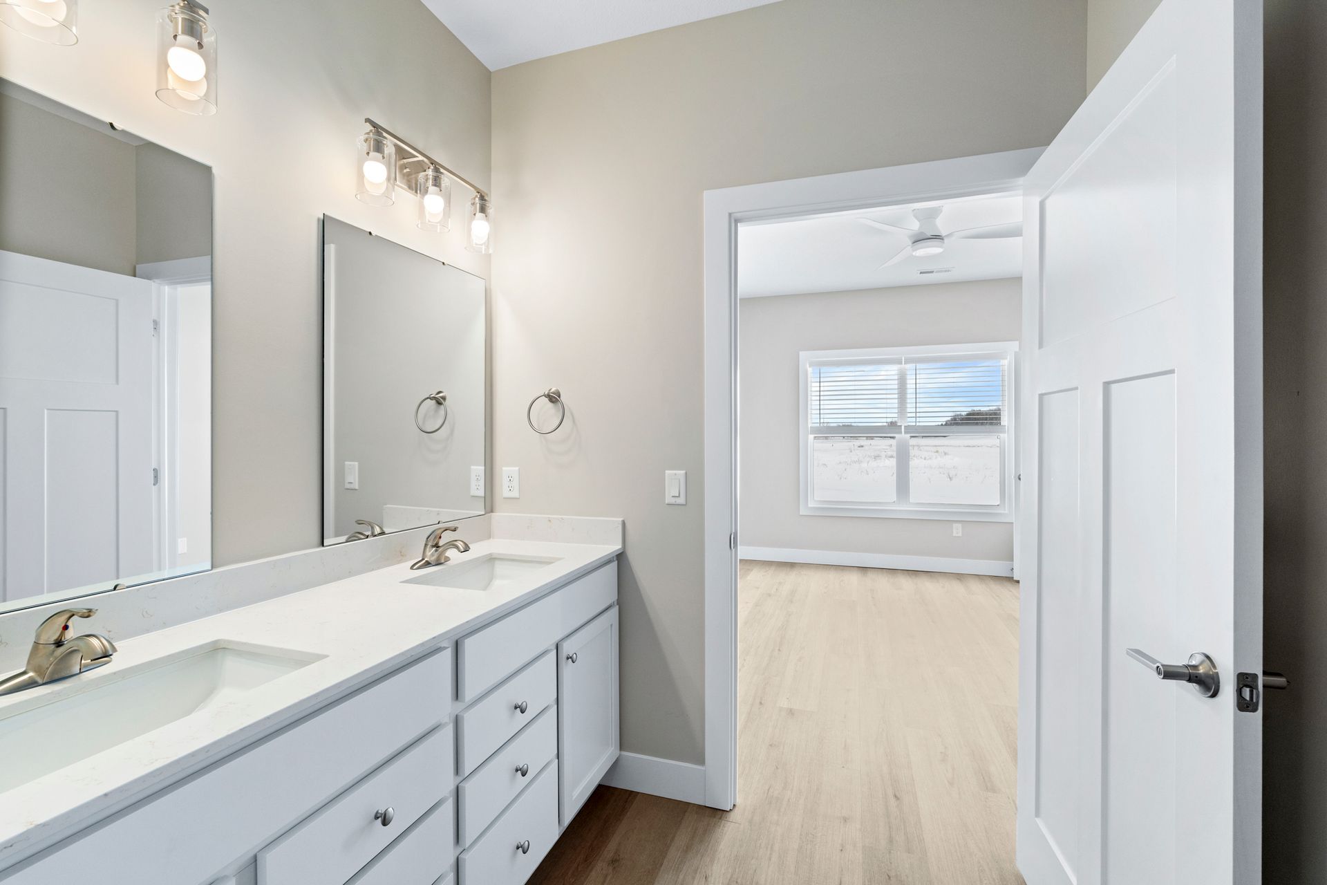Bathroom with double sinks, white cabinets, and a doorway leading to a room with a window and wood flooring.