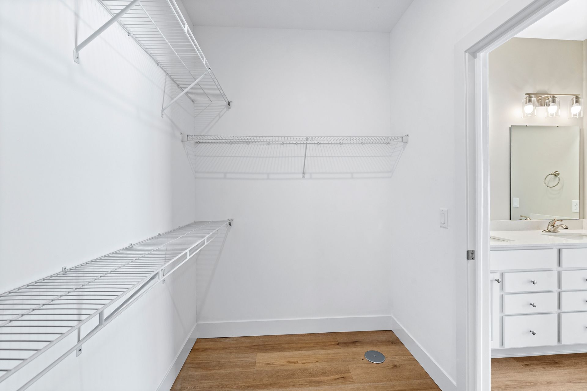 Empty white closet with wire shelving and wooden floor, doorway to bathroom.