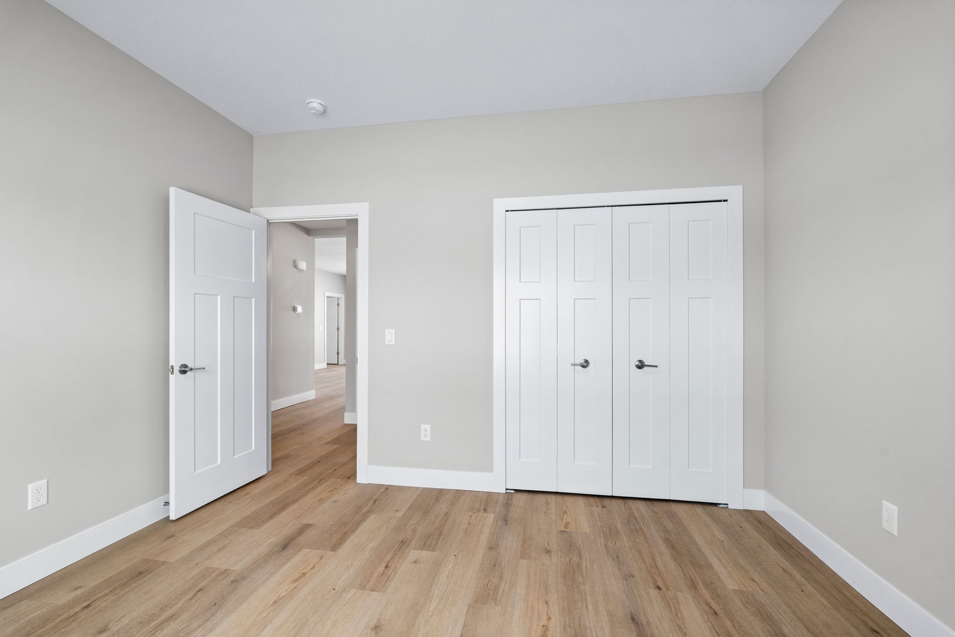 Empty bedroom with light wood floor, white doors, and light gray walls.