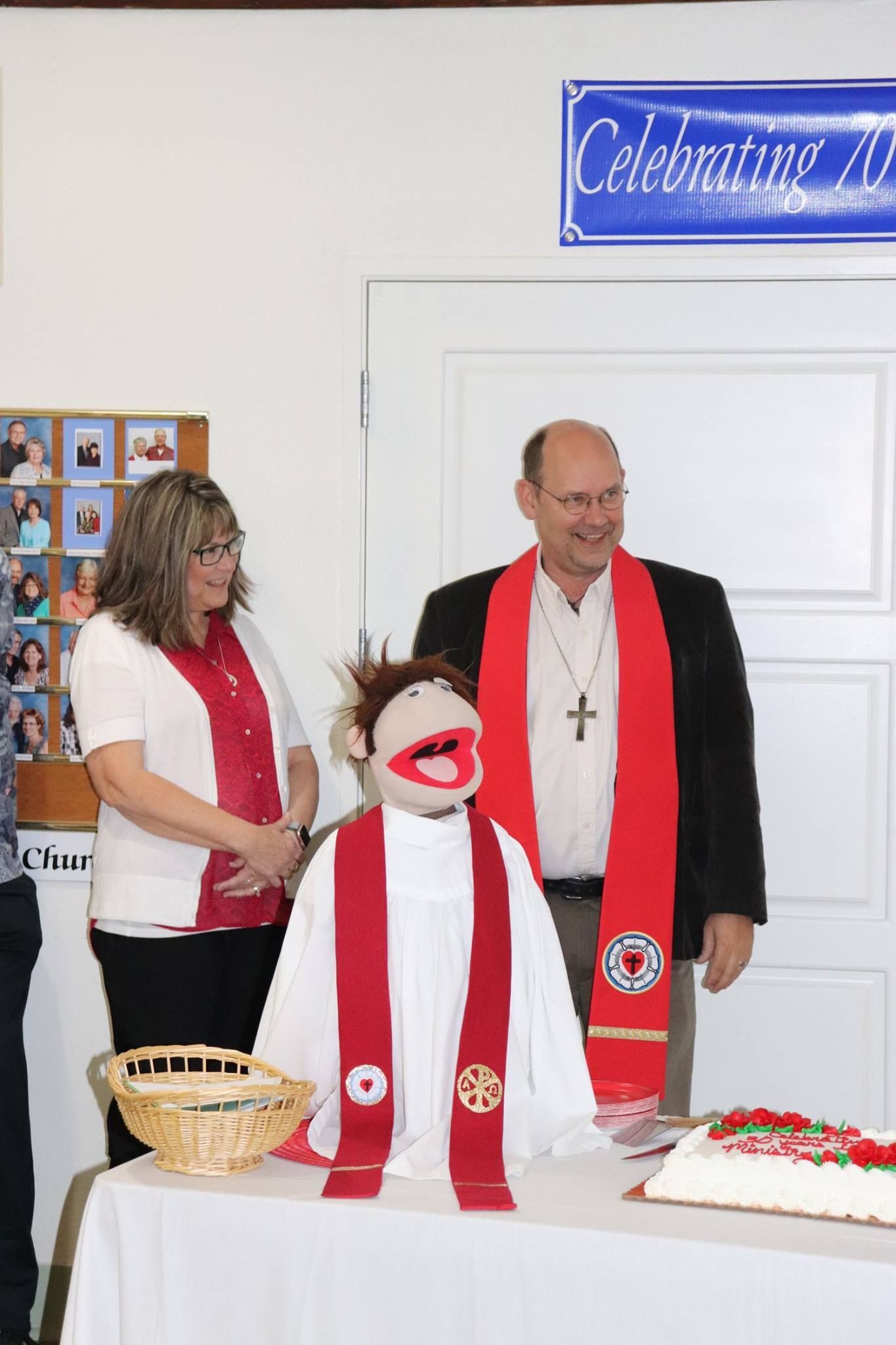 Three people with a puppet at a celebration. Woman, man, and puppet wearing red stoles stand by a cake.