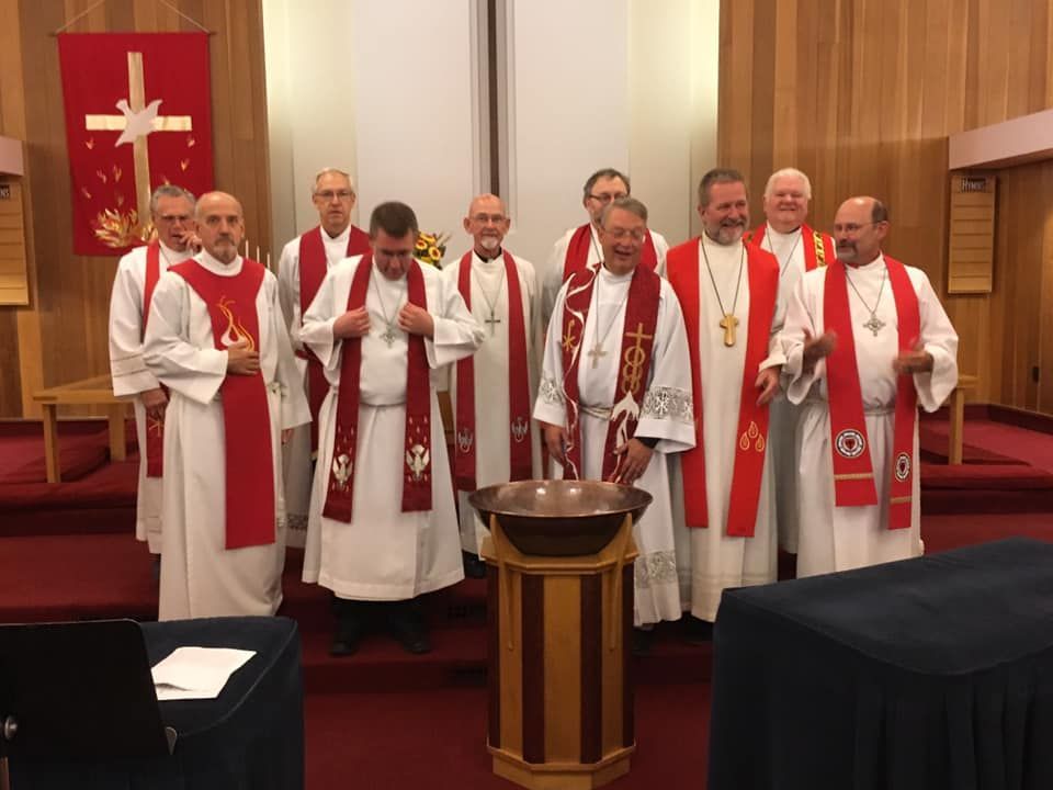 Clergy in red stoles gather around a baptismal font in a church, smiling, and looking at the camera.