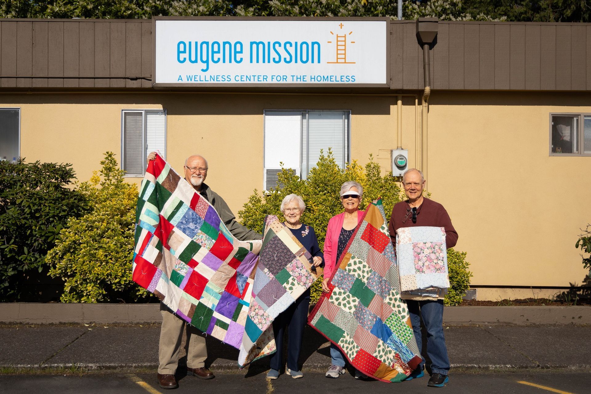 Four people hold quilts outside Eugene Mission building.
