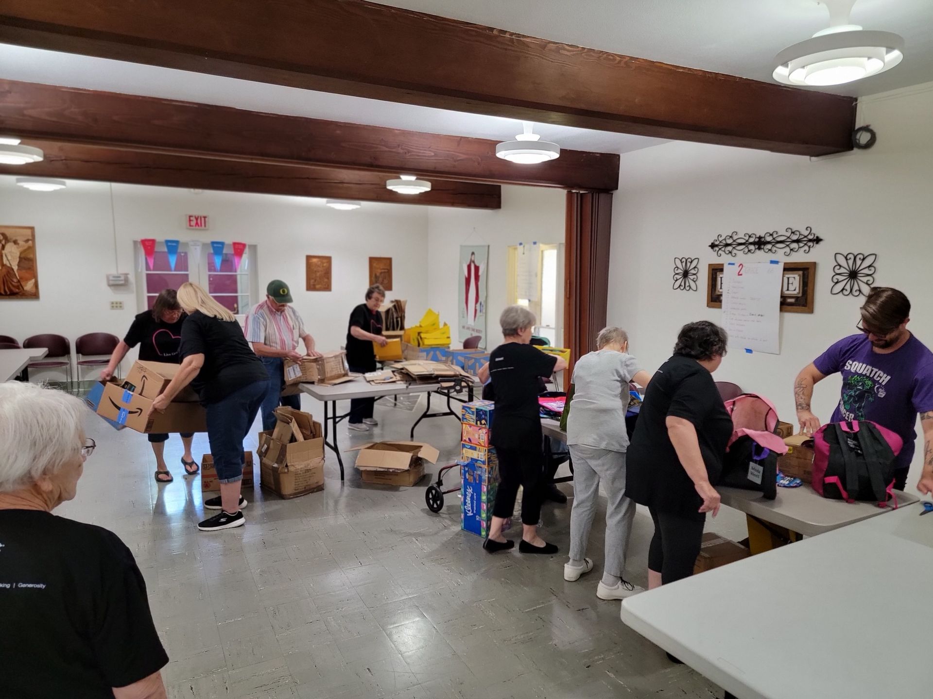 People packing boxes and sorting items in a community room. Tables are covered with boxes and supplies.