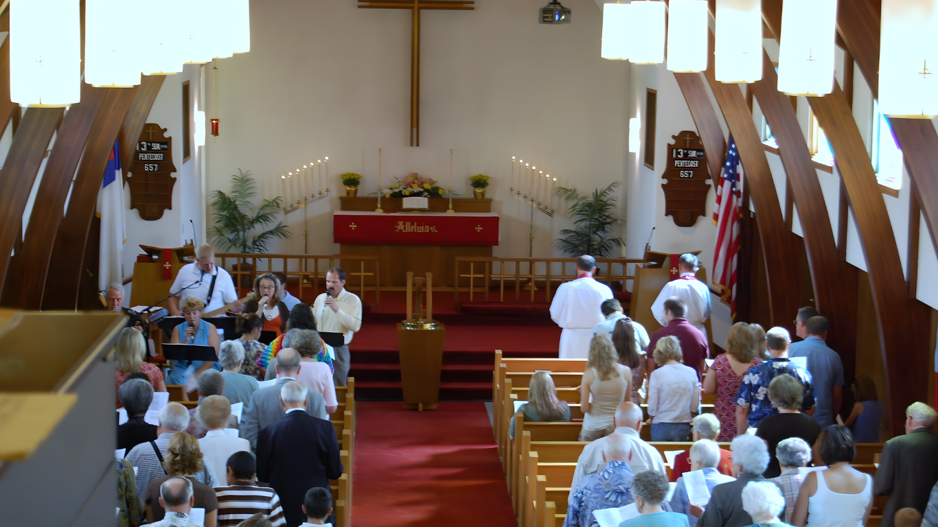 Interior church view with rows of people in pews, red carpet aisle, and a cross.