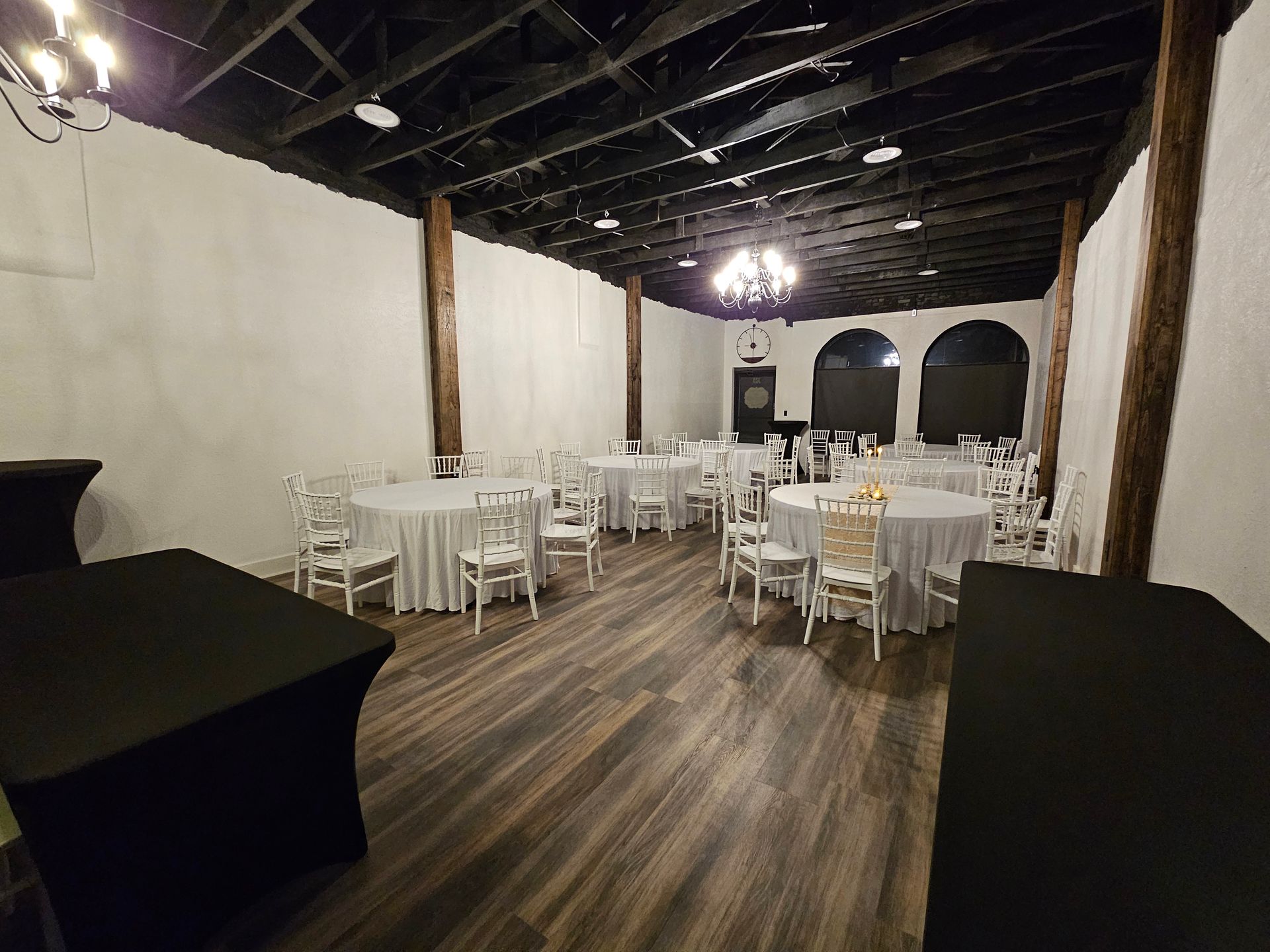 Several round tables covered in white tablecloths surrounded by white Chiavari chairs in a venue with wood-look flooring.