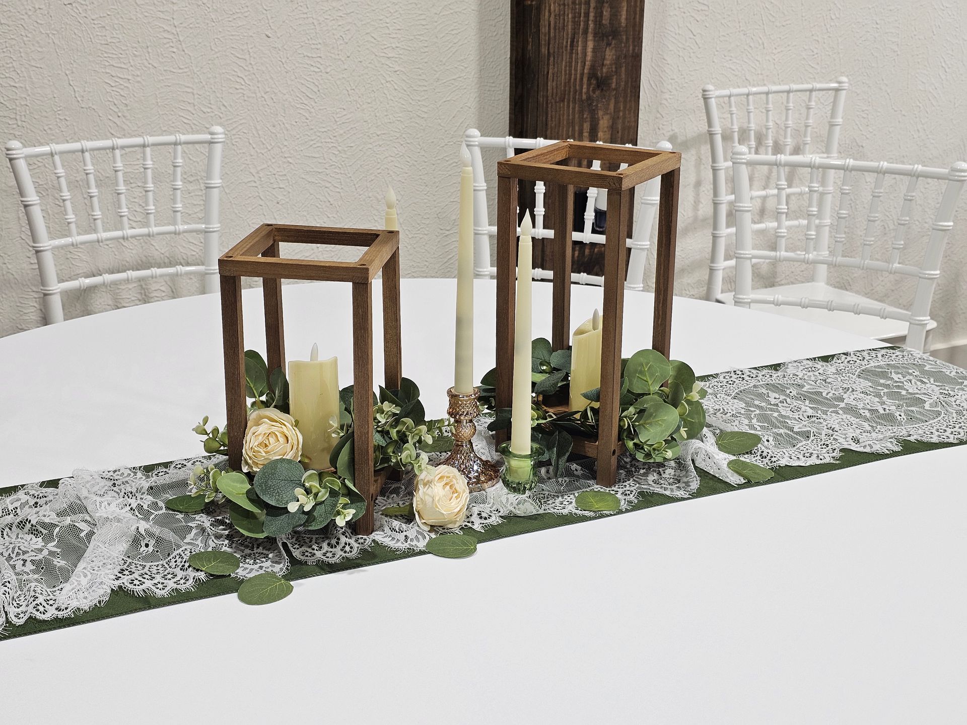 A rustic wedding centerpiece with wooden lantern frames, taper candles, and greenery on a lace runner on a white table.