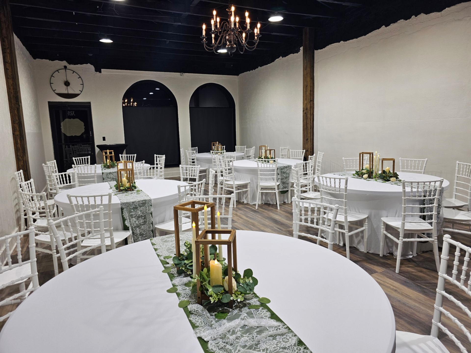 Several round tables covered in white tablecloths surrounded by white Chiavari chairs in a venue with wood-look flooring.