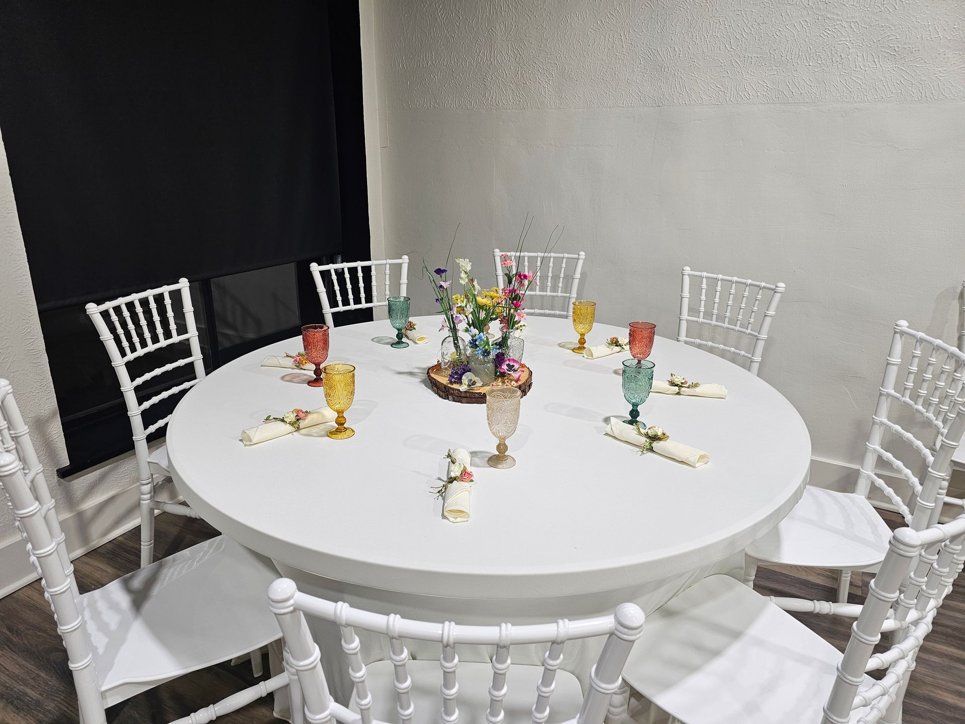 Several round tables covered in white tablecloths surrounded by white Chiavari chairs in a venue with wood-look flooring.