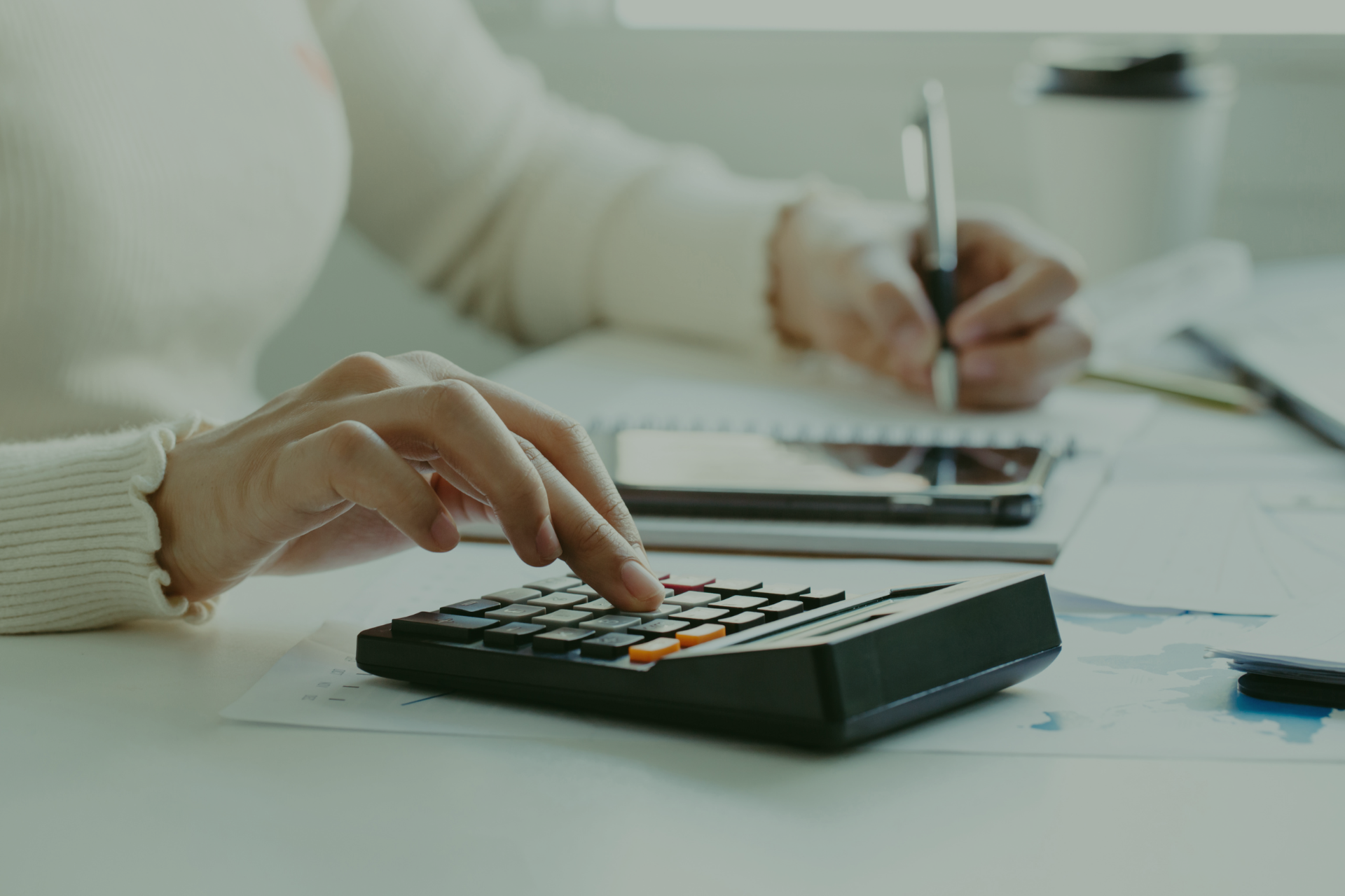Person using a calculator and writing in a notebook at a desk.
