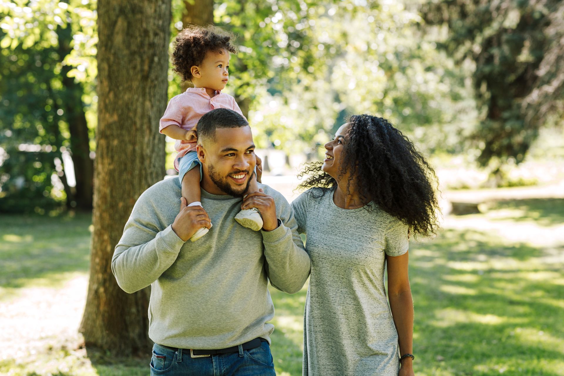 Family in park: Dad carries child on shoulders, Mom smiles. Green trees and sunny day.