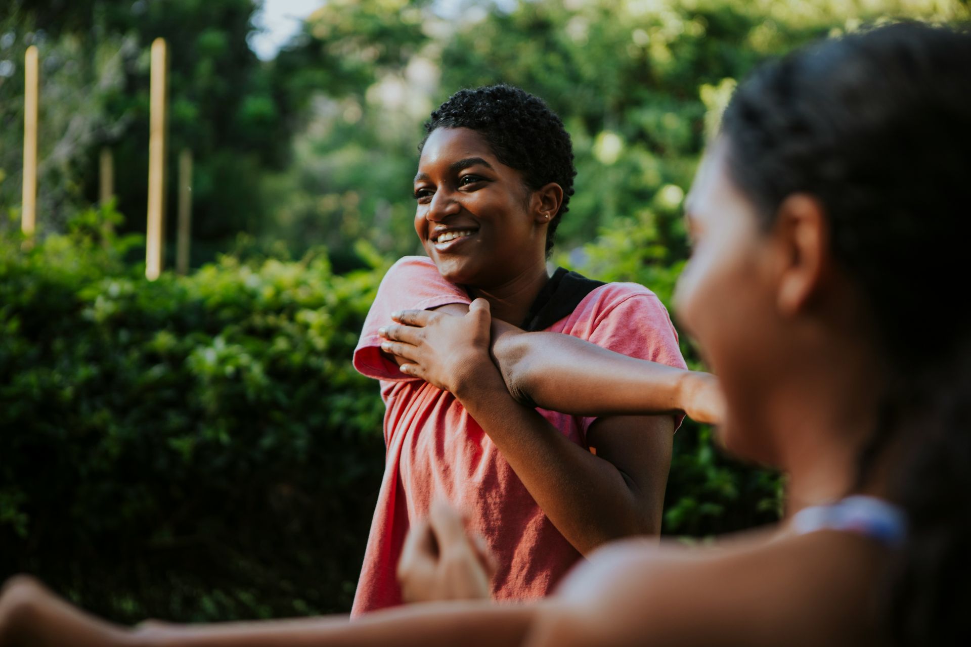 Two women stretching outdoors, one smiles, with a green backdrop.