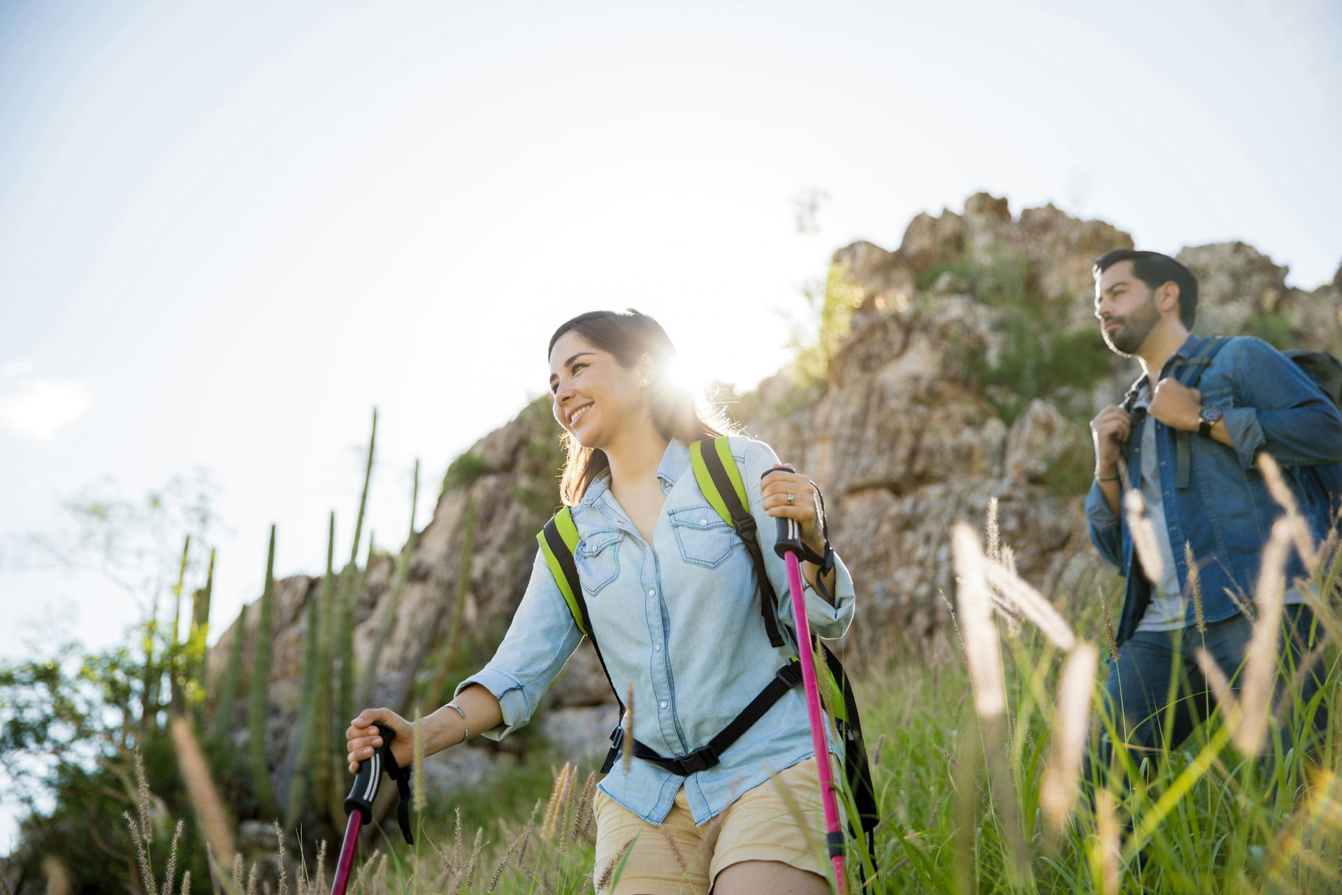 A woman and man hike up a sunny trail with backpacks and poles.