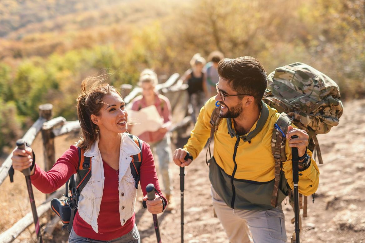 Group hikes up a trail. A smiling couple leads, using poles, in autumn sunlight. Others follow.