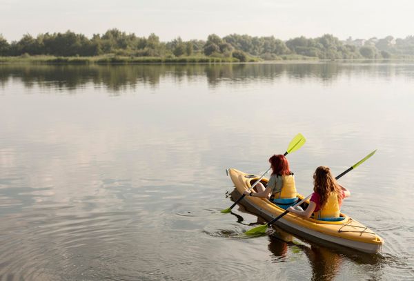 Two people kayaking on a calm lake with trees in the background.
