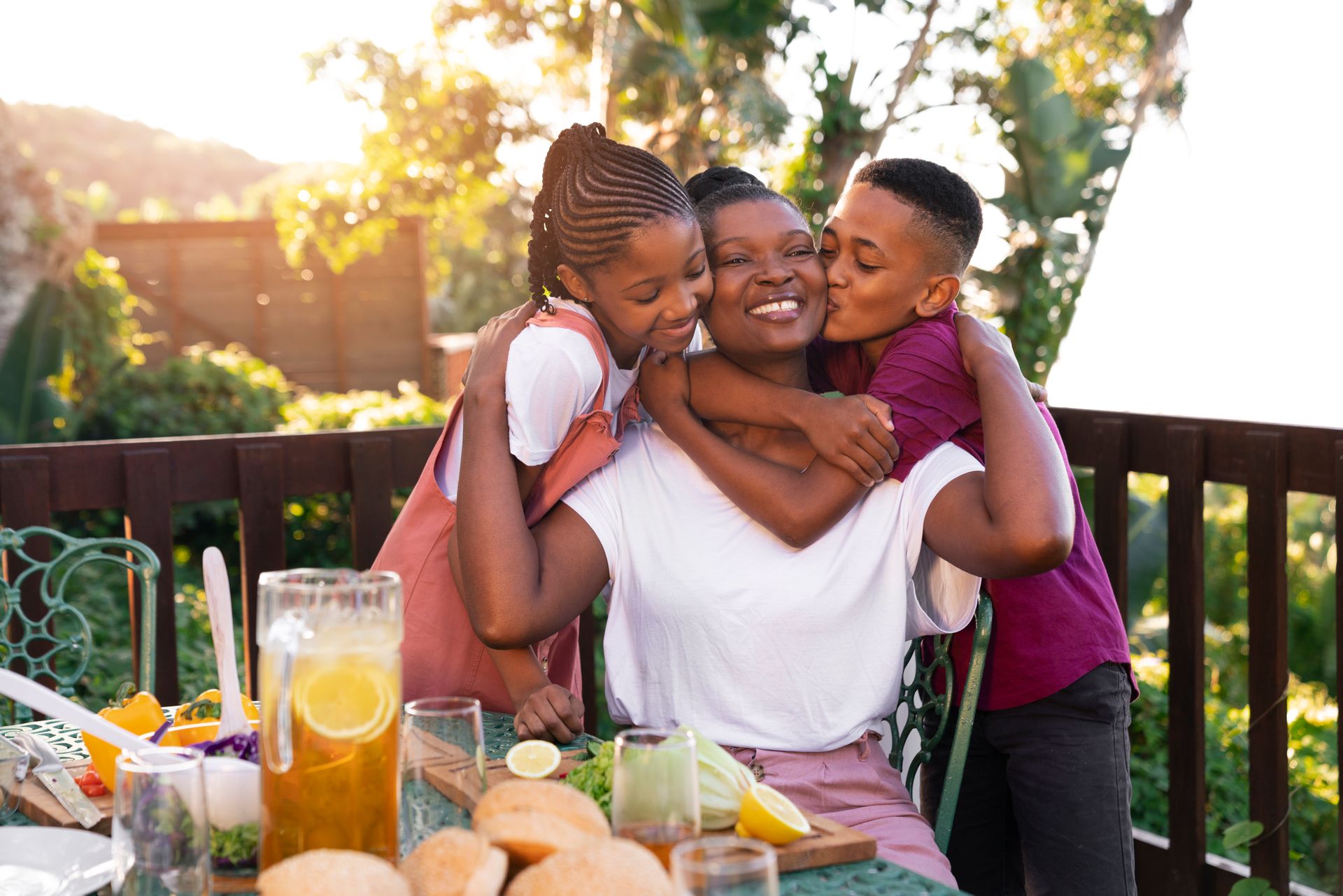 Mother embraces children, receiving hugs and kisses at an outdoor table.