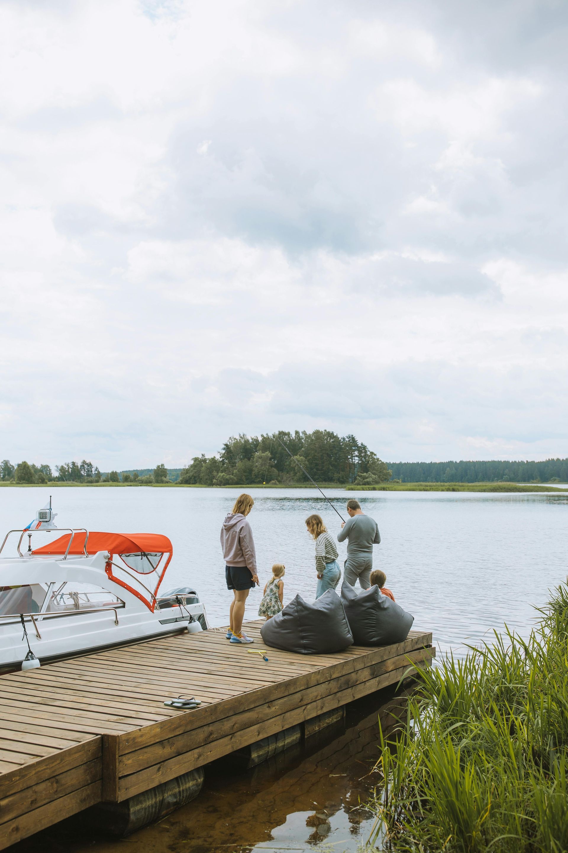 Family on a wooden dock by a lake, cloudy sky. People are sitting on beanbags near a boat.