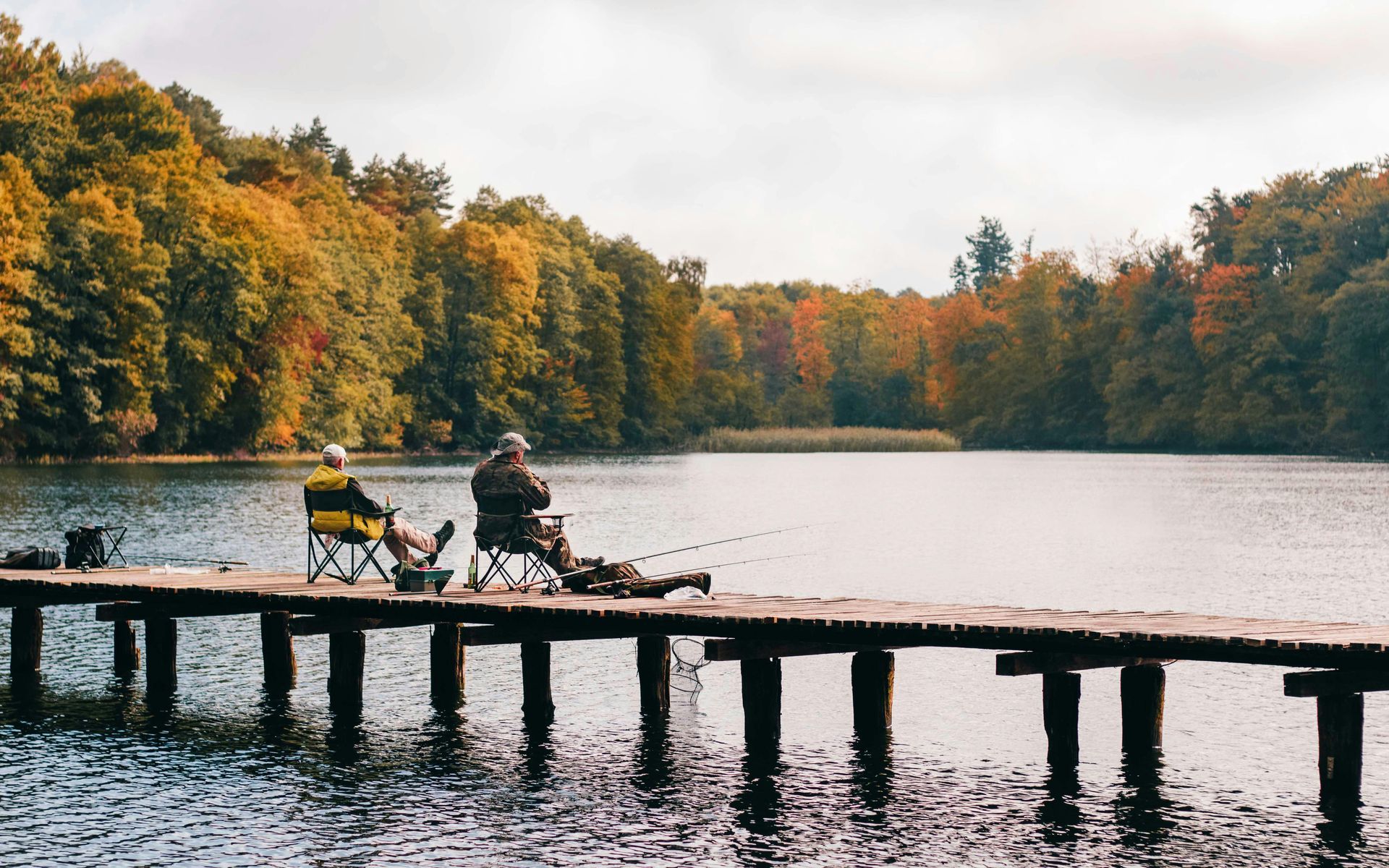 Two people fishing from a wooden pier on a lake surrounded by fall trees.